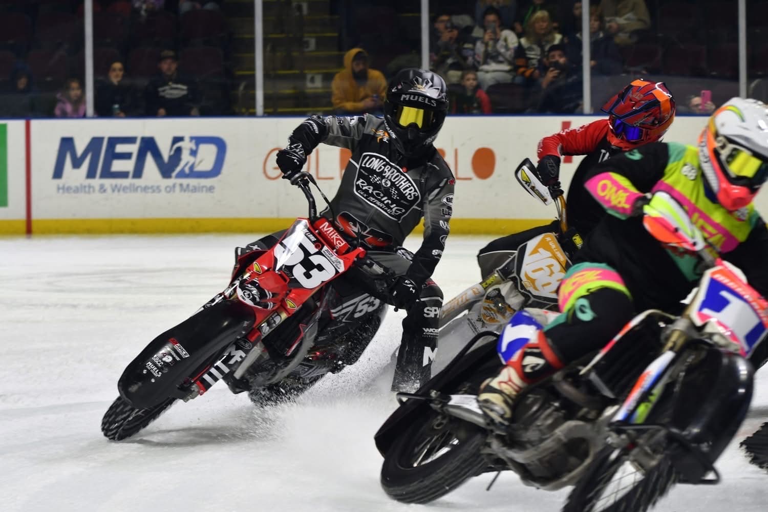 Three flat track motorcycles leaning into a corner together on arena ice