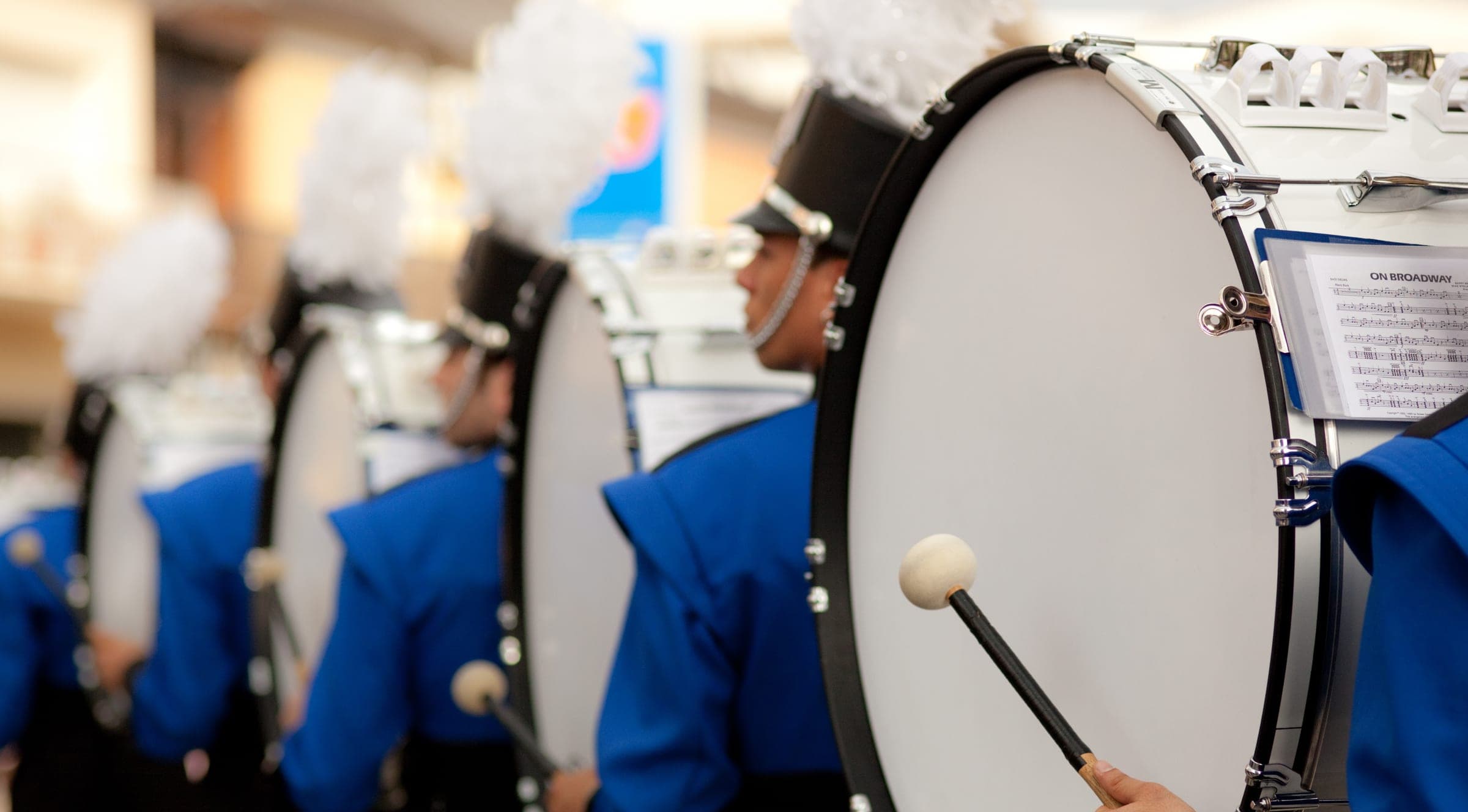 A marching band drum line in blue uniforms and plumed shakos, bass drums in a row